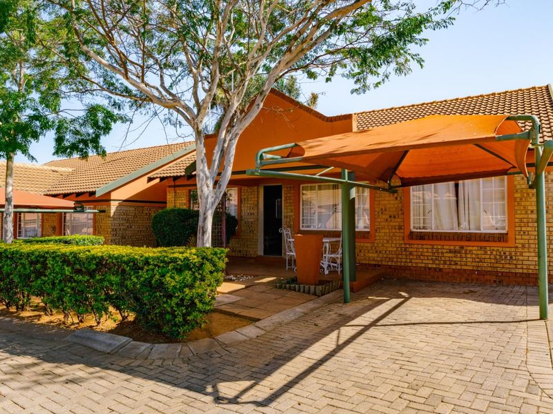 Front view of a townhouse featuring a brick facade, tiled roof, paved driveway, and a covered patio area with established garden landscaping.