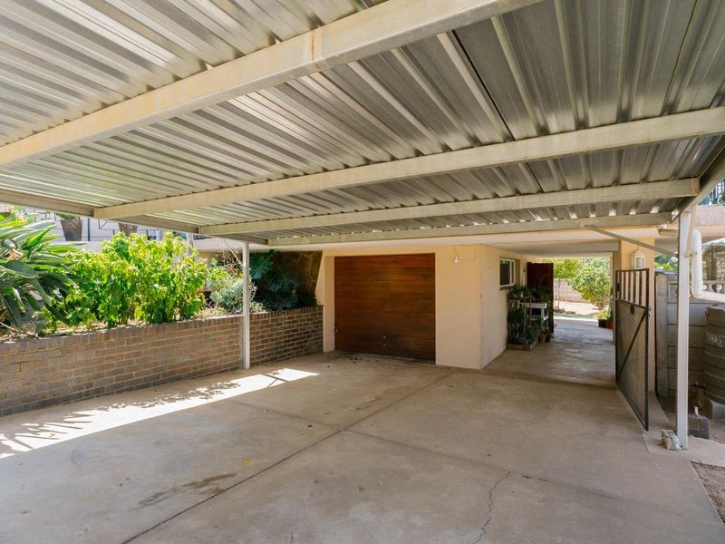 A covered carport area featuring a corrugated metal roof, concrete flooring, and a secure wooden garage door with adjacent brick retaining wall.