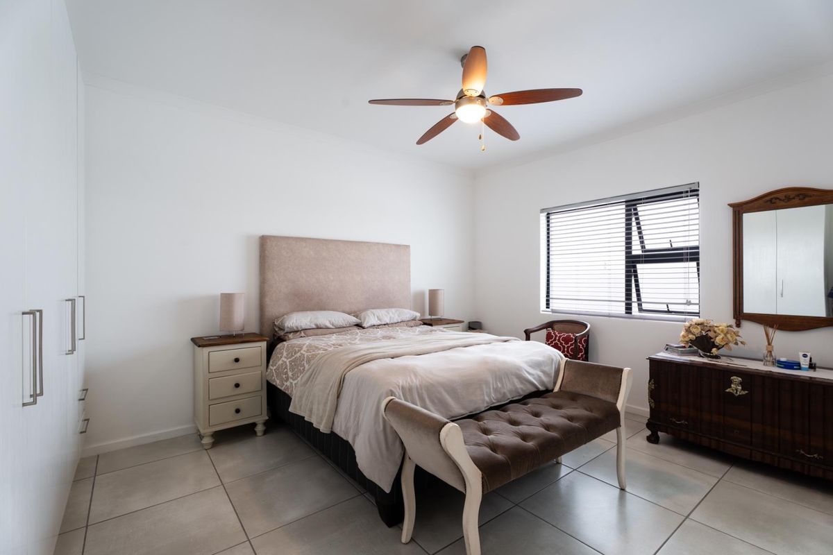 A well-maintained bedroom featuring neutral tiled flooring, built-in cupboards, a ceiling fan, and large windows with venetian blinds.