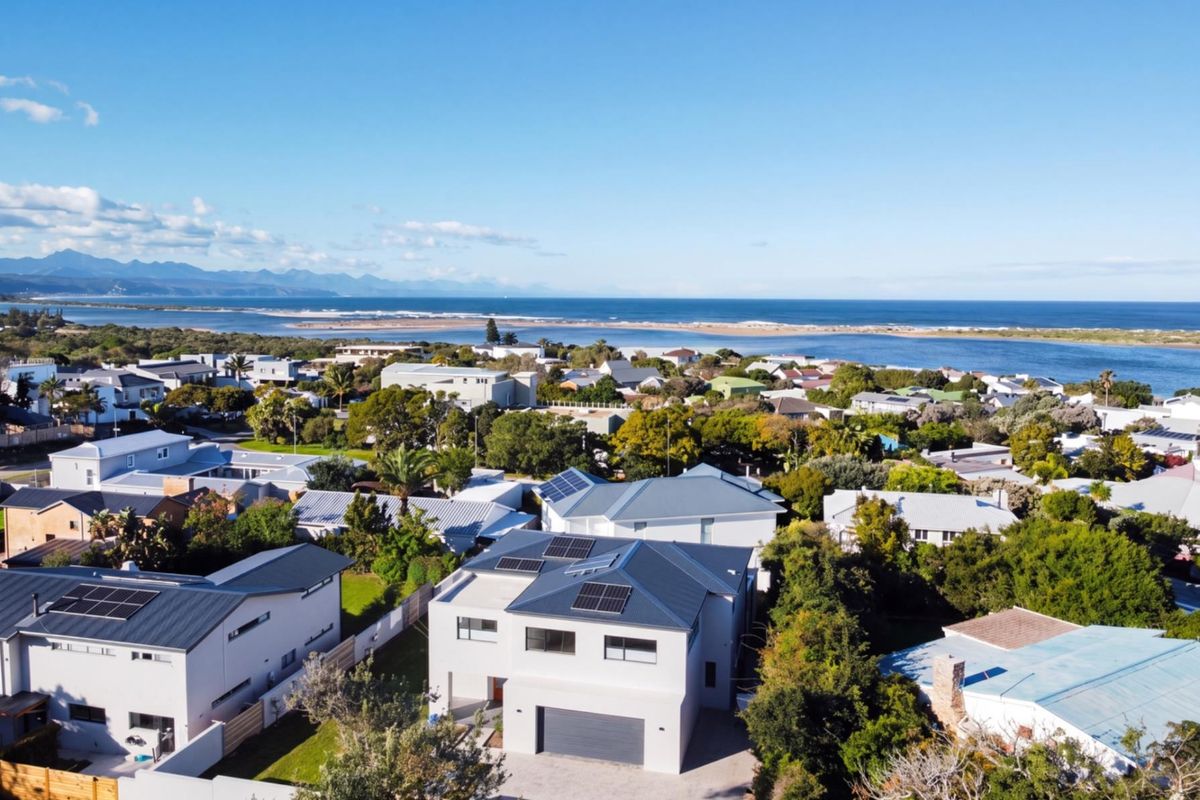 Aerial view of a modern residential property featuring a metal roof with solar panels, a paved driveway, and proximity to the coastline.