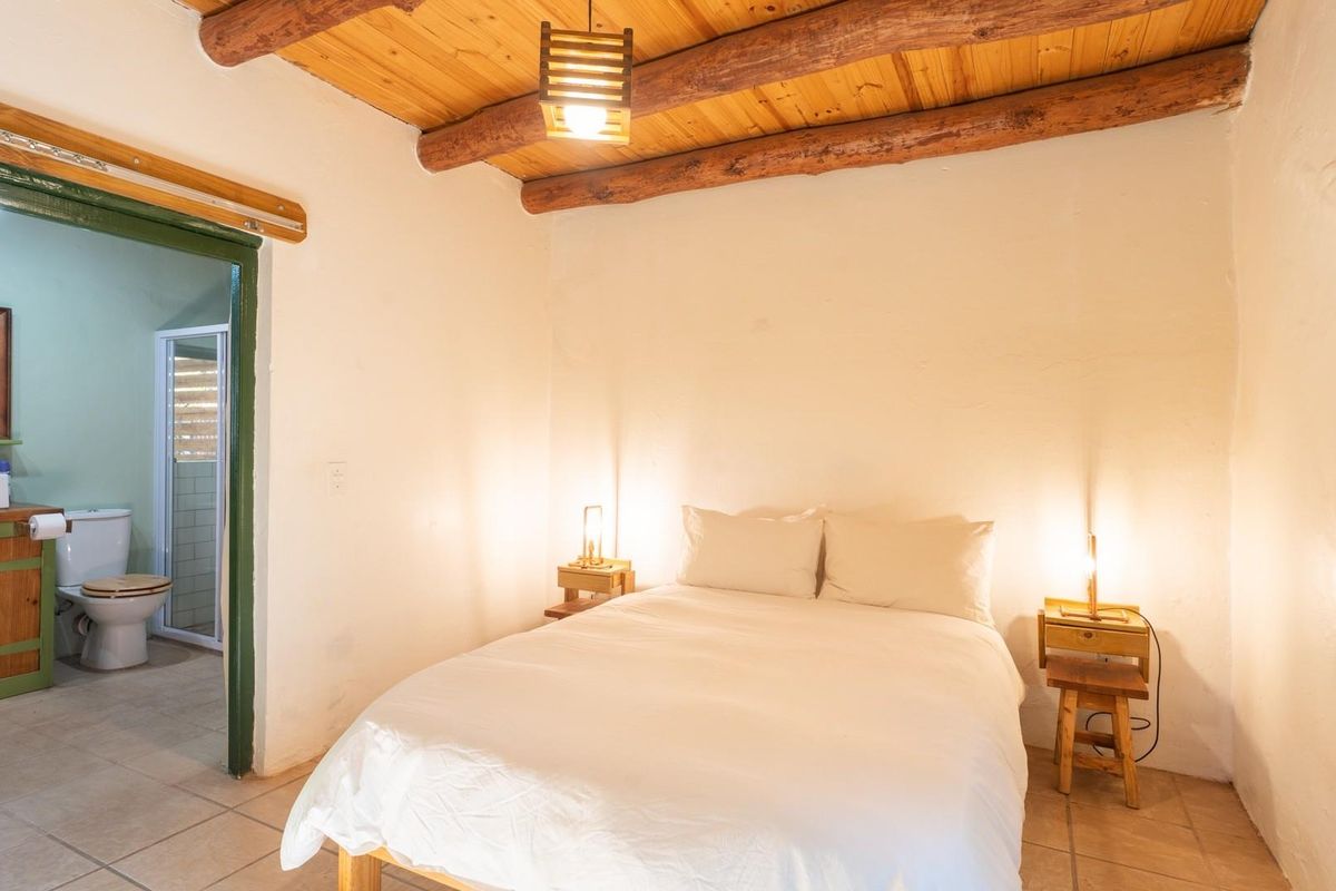 A rustic bedroom featuring a timber beam ceiling, tiled flooring, and an adjoining en-suite bathroom with a wooden vanity.