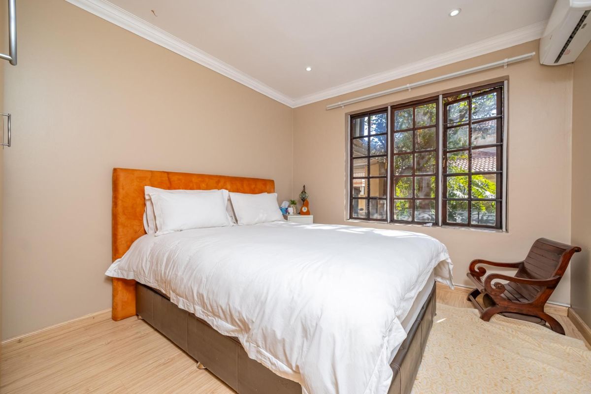 A well-maintained bedroom featuring light wood-look flooring, crown molding, a wall-mounted air conditioning unit, and large multi-pane windows.