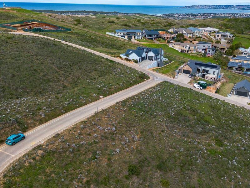 An aerial view of a residential estate featuring vacant land plots, paved roads, and established modern homes with coastal views in the distance.