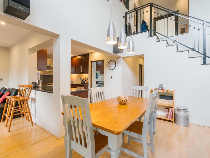 Open plan living and dining area featuring a double-volume ceiling, staircase with wrought iron balustrade, and an adjacent kitchen with dark cabinetry.