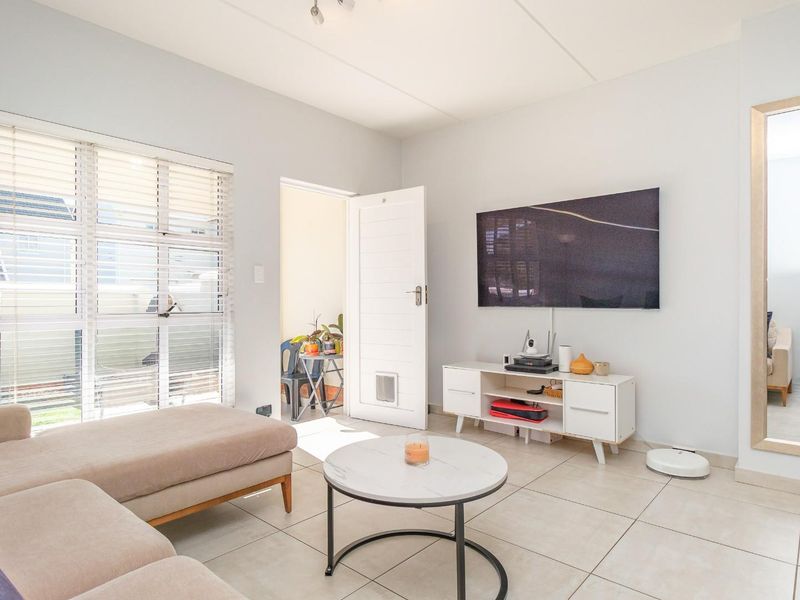 A bright and well-maintained living room featuring neutral tiled flooring, large windows with venetian blinds, and a clean, modern aesthetic.
