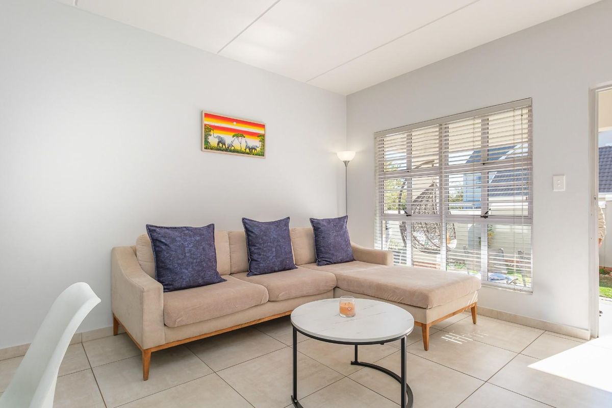 A bright and well-maintained living room featuring neutral tiled flooring, large windows with venetian blinds, and direct access to an outdoor patio area.