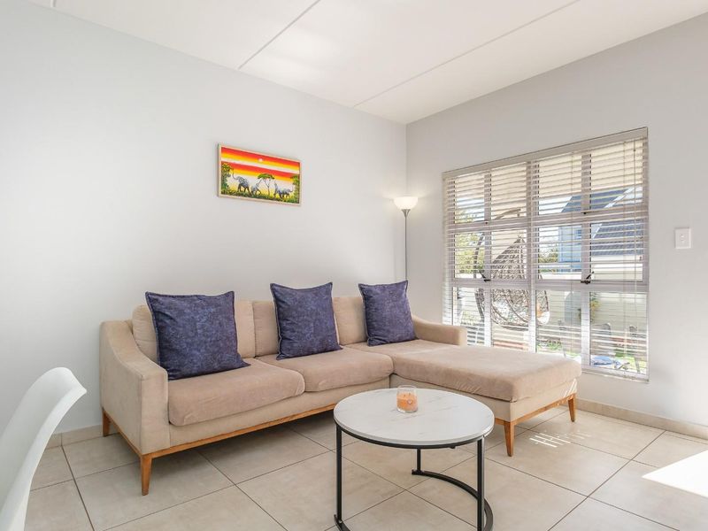 A bright and well-maintained living room featuring neutral tiled flooring, large windows with venetian blinds, and direct access to an outdoor patio area.