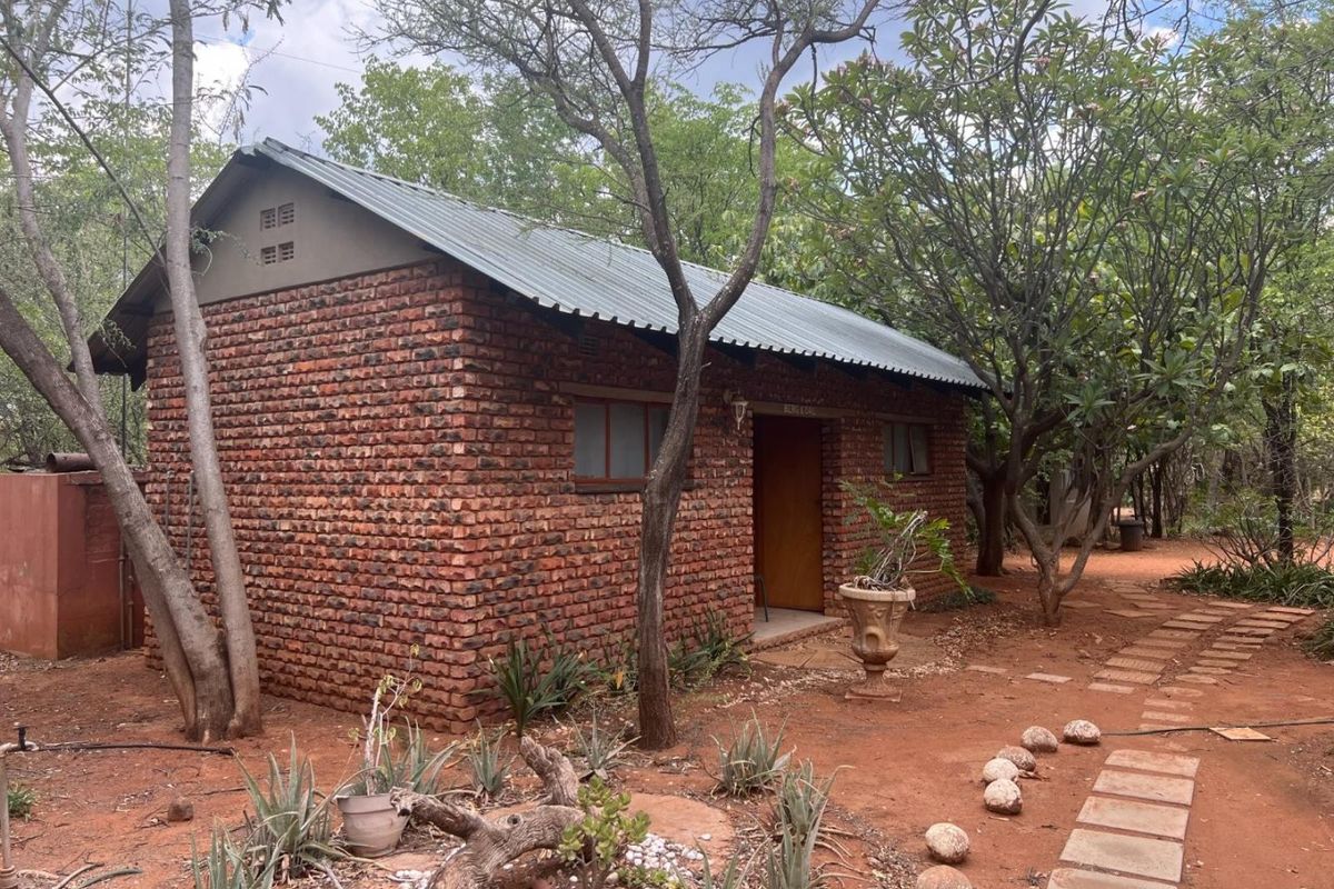 A rustic face-brick cottage set within a natural bushveld garden, featuring a pitched metal roof and established surrounding trees.