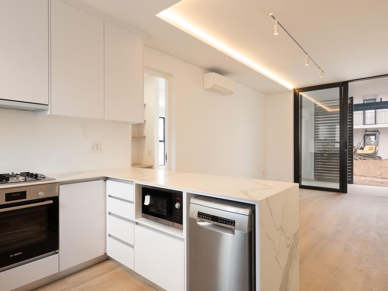 Modern open-plan kitchen featuring sleek white cabinetry, stone countertops, integrated appliances, and timber flooring, flowing seamlessly to an outdoor patio area.