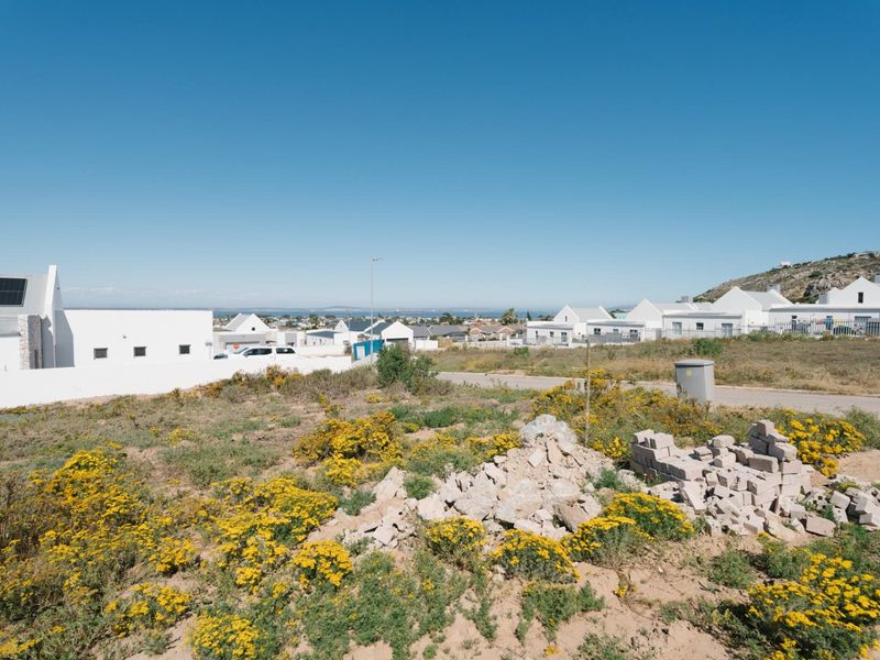 A vacant residential plot featuring natural vegetation and a utility box, situated in a developed coastal neighborhood with surrounding residential properties.