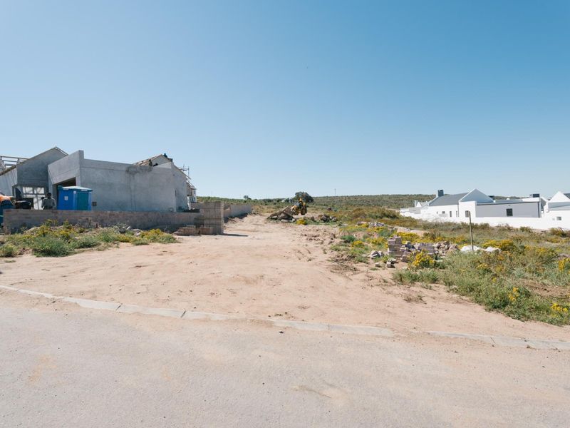 A vacant residential plot under development, featuring an unpaved access area and ongoing construction of a neighboring structure.