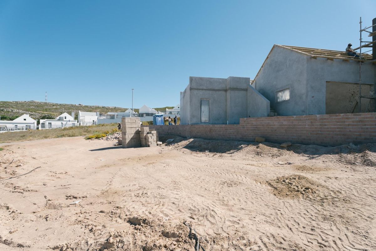 A view of a residential property under construction, featuring an unfinished building structure, roof trusses, and a brick boundary wall on a sandy plot.