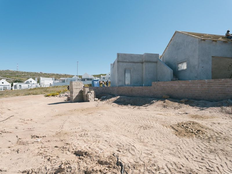 A view of a residential property under construction, featuring an unfinished building structure, roof trusses, and a brick boundary wall on a sandy plot.