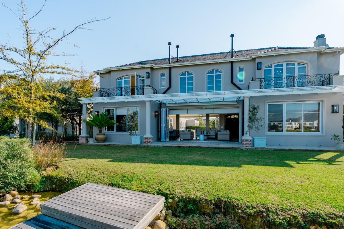 Rear view of a luxury two-story residence featuring a covered patio, upper-level balconies with wrought iron railings, and a landscaped garden with a private jetty.