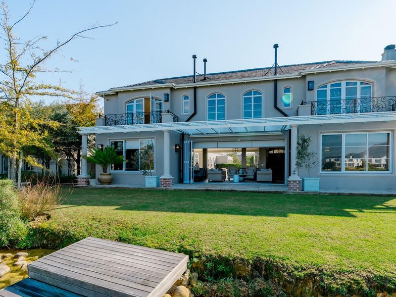 Rear view of a luxury two-story residence featuring a covered patio, upper-level balconies with wrought iron railings, and a landscaped garden with a private jetty.