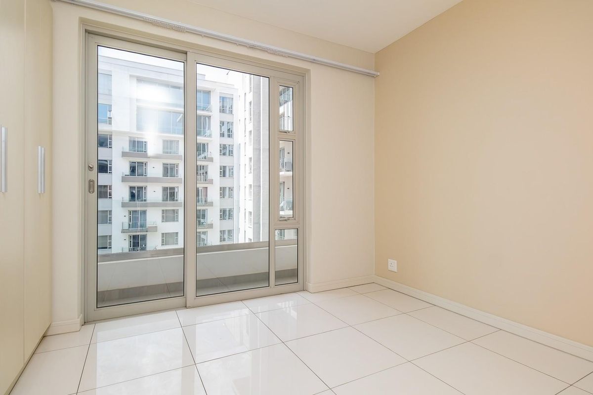 A bright, empty bedroom featuring neutral tiled flooring, built-in cupboards, and a large sliding glass door providing access to a balcony.
