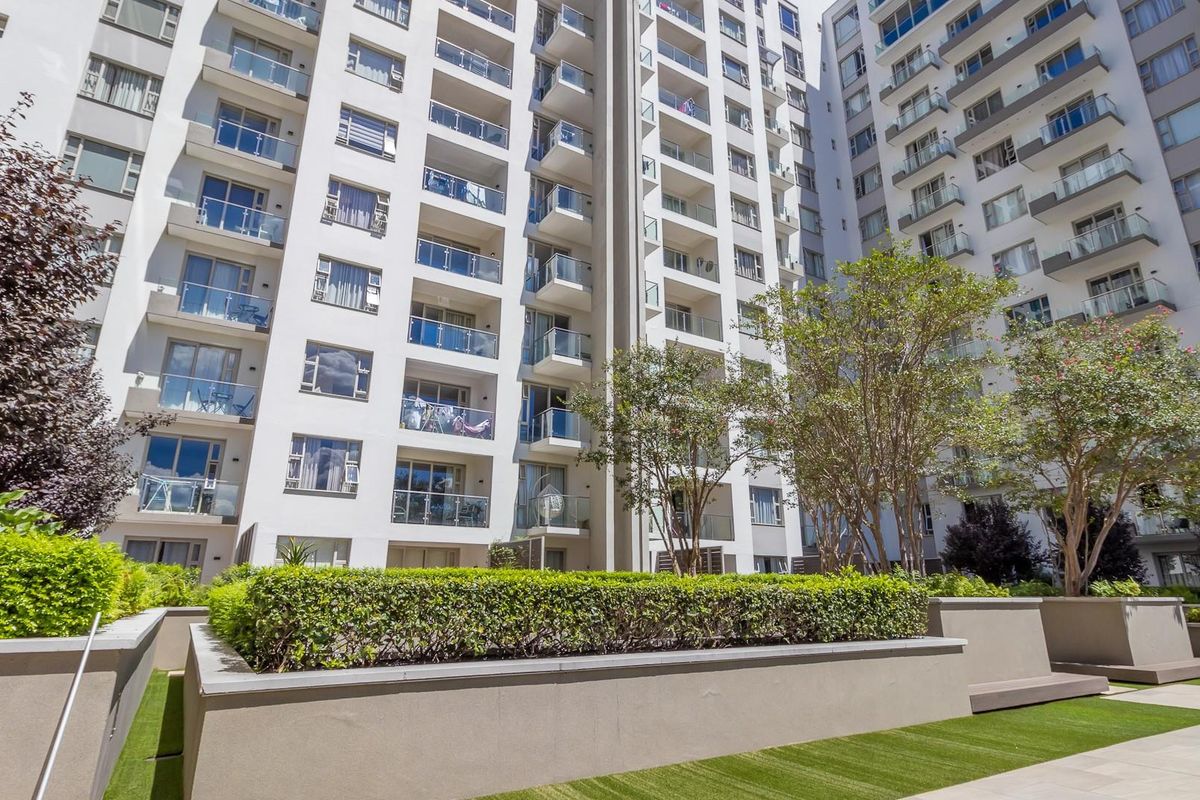 A view of the apartment complex rear facade featuring multiple balconies with glass balustrades, overlooking a well-maintained landscaped garden area.