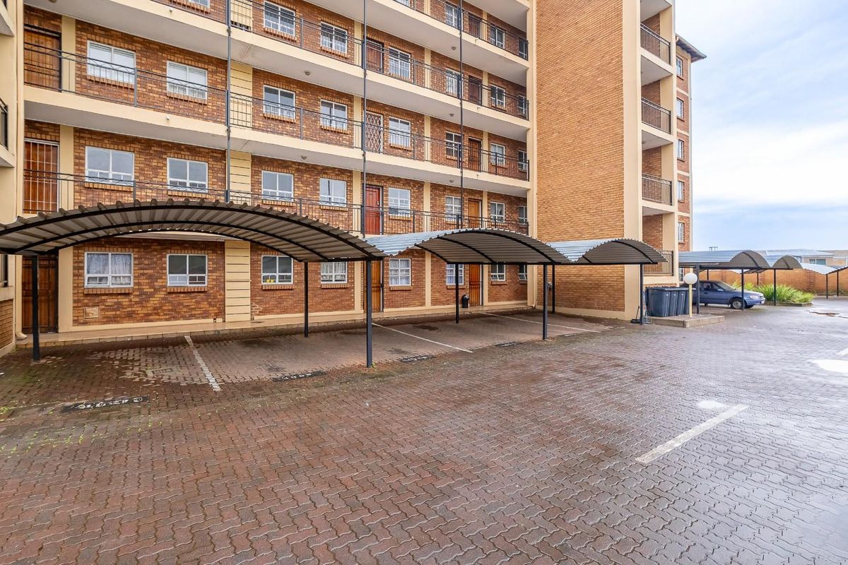 Exterior view of an apartment complex featuring a brick facade, multiple balconies with metal railings, and a paved parking area with covered bays.