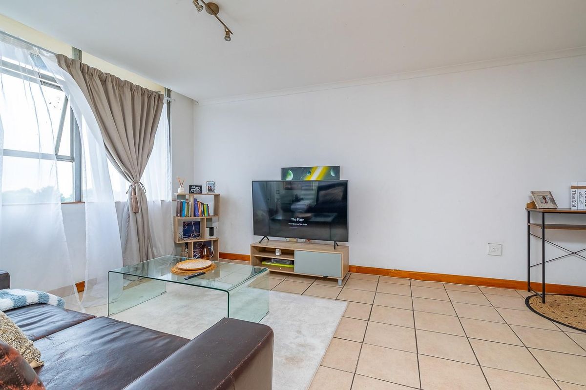 A bright living room featuring neutral tiled flooring, large windows with natural light, and clean wall finishes.