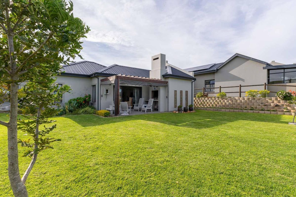 A well-maintained rear garden featuring a spacious lawn, a covered patio with a built-in braai, and a tiered retaining wall.