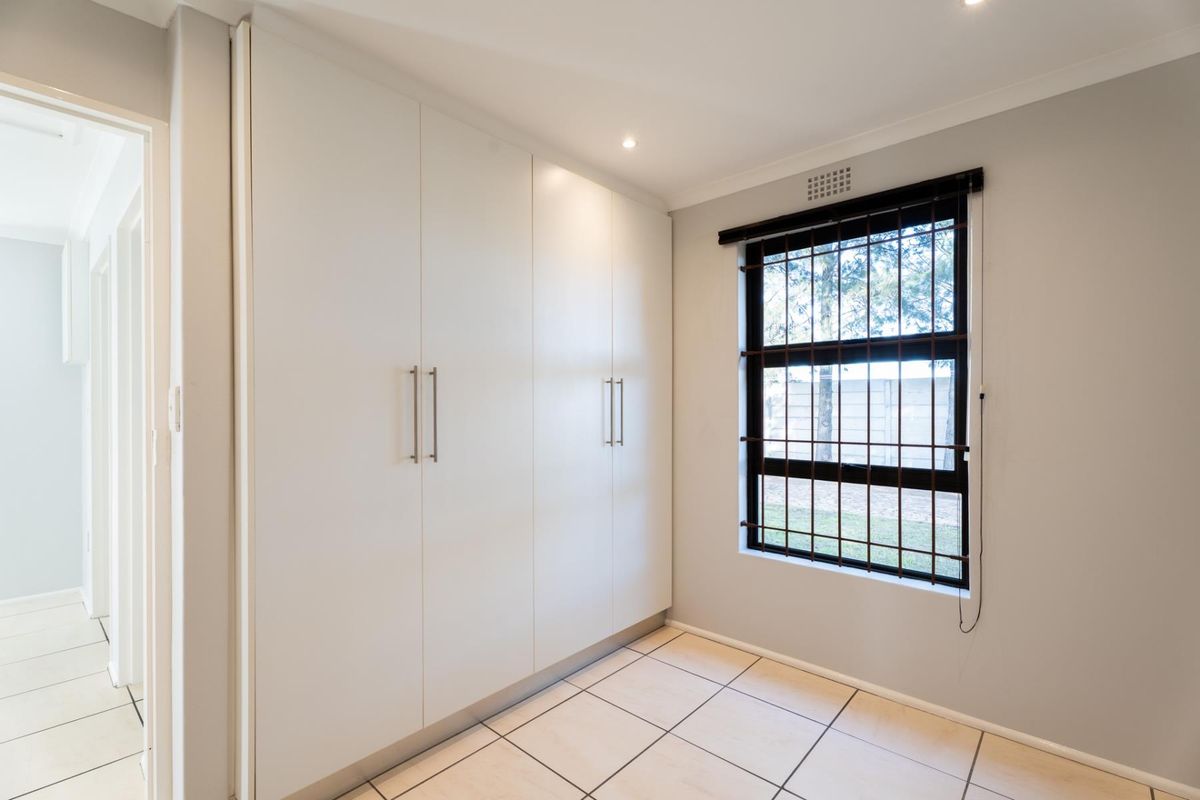 A clean, empty bedroom featuring neutral tiled flooring, ample built-in cupboard storage, and a window fitted with security bars.