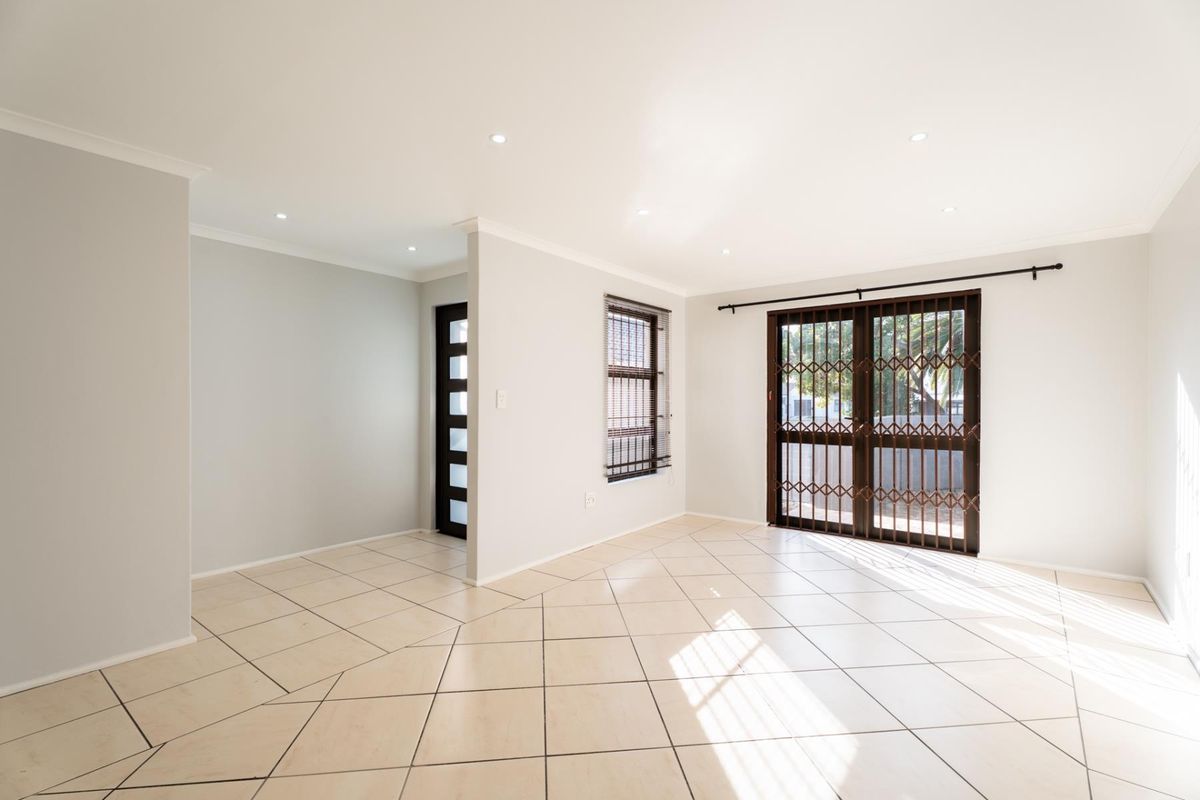 A bright, open-plan living area featuring neutral tiled flooring, recessed downlights, and a sliding glass door with a security gate leading outdoors.