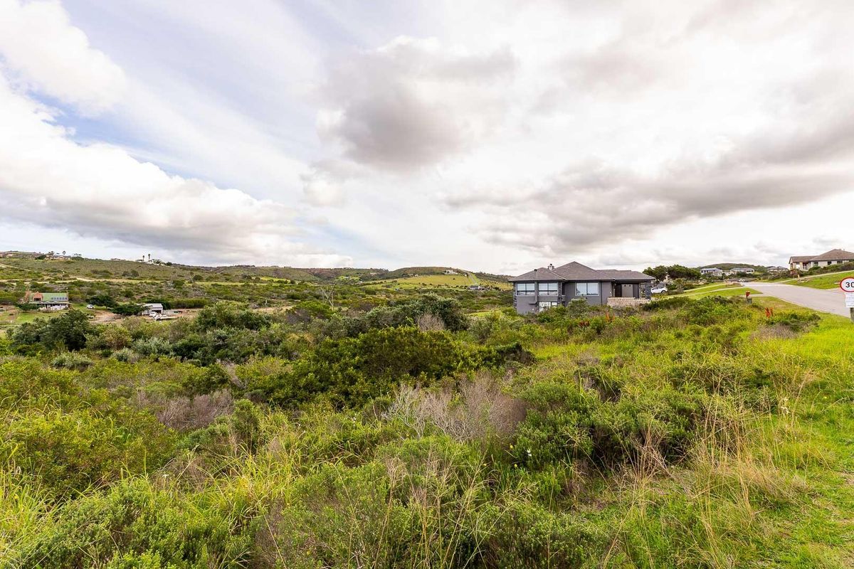 A wide-angle view of a vacant residential plot featuring natural landscape, surrounding neighborhood infrastructure, and a paved access road.