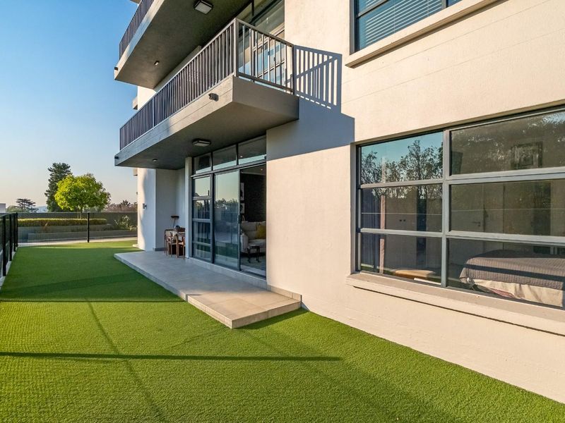Rear view of the apartment building featuring a ground-level patio with artificial turf, sliding glass doors, and an upper-level balcony.