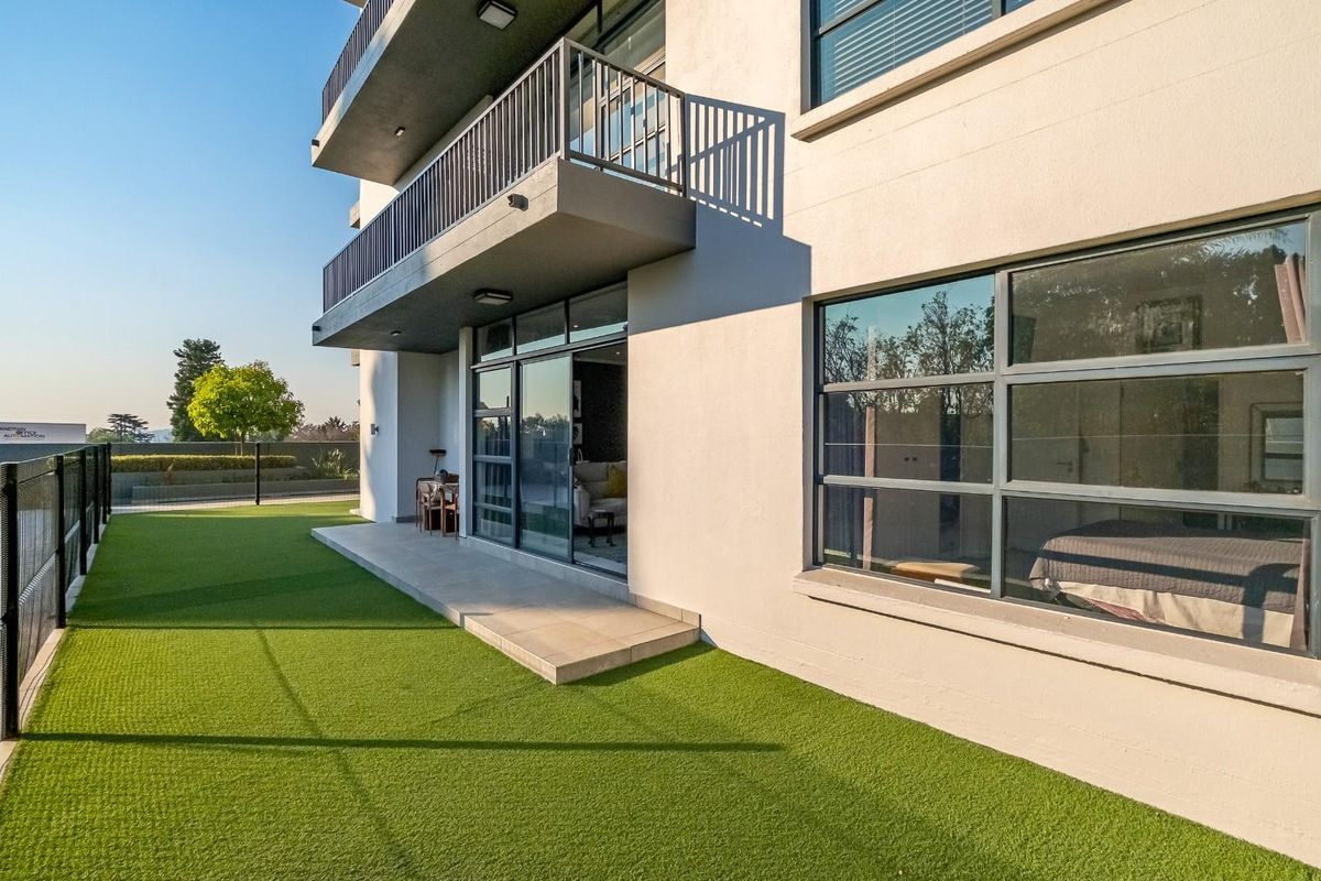 Rear view of the apartment building featuring a ground-level patio with artificial turf, sliding glass doors, and an upper-level balcony.
