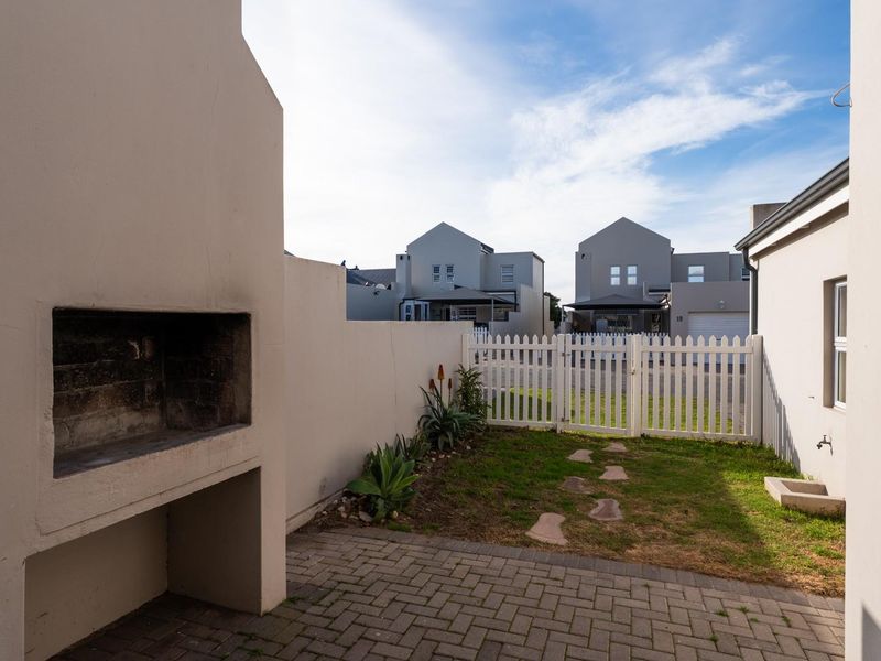 A private rear courtyard featuring a paved patio area with a built-in braai, small lawn, and white picket fence enclosure.