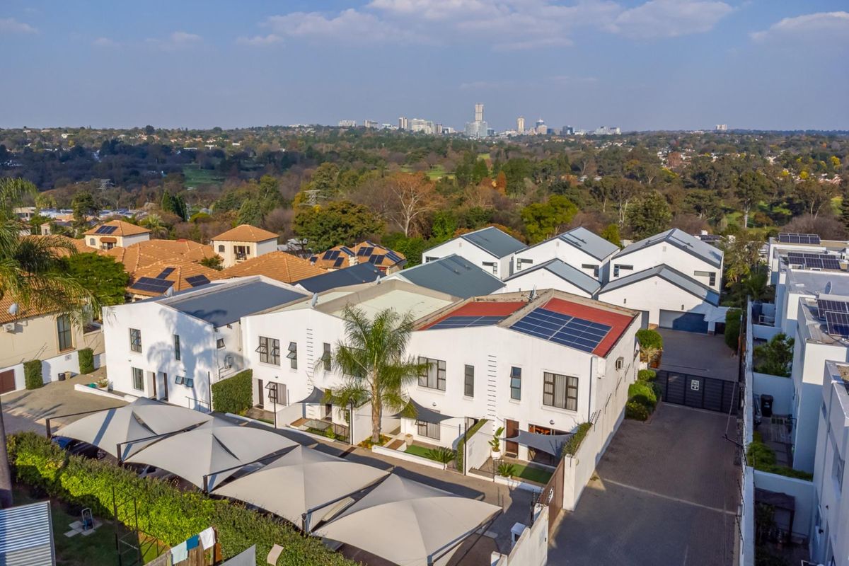 Aerial view of a residential complex featuring multiple units with solar panels, paved driveways, and covered parking areas set against a city skyline.