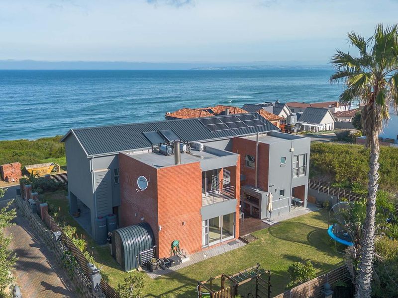 Aerial view of a modern multi-level coastal home featuring brick and grey finishes, solar installations, and a spacious rear garden overlooking the ocean.