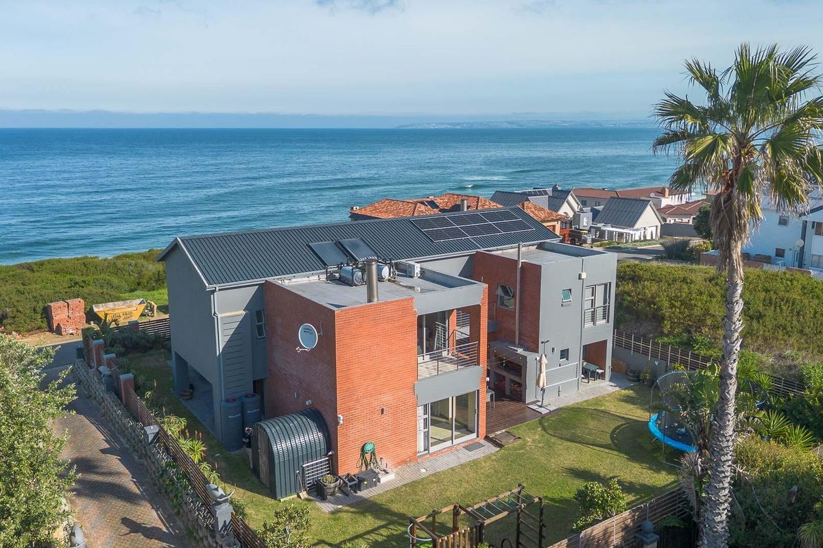 Aerial view of a modern multi-level coastal home featuring brick and grey finishes, solar installations, and a spacious rear garden overlooking the ocean.