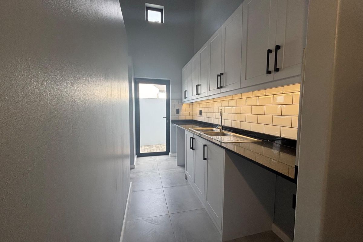 A modern, clean scullery featuring white cabinetry, dark granite countertops, subway tile backsplash, and under-cabinet lighting, leading to an exterior door.