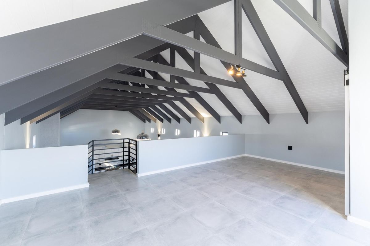 Spacious open-plan loft area featuring high vaulted ceilings with exposed dark timber trusses, modern tiled flooring, and a sleek metal staircase balustrade.