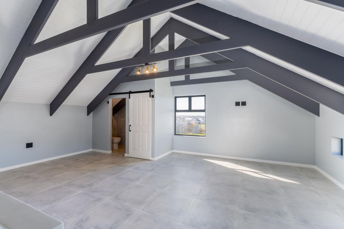 Spacious attic bedroom featuring vaulted ceilings with exposed dark timber trusses, tiled flooring, and a sliding barn door leading to an en-suite bathroom.