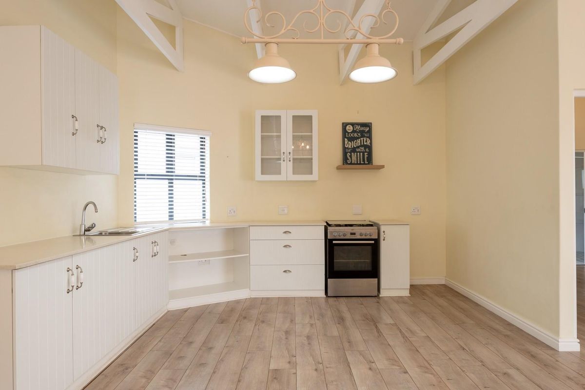A bright, open-plan kitchen featuring white cabinetry, laminate countertops, a freestanding oven, and exposed ceiling trusses with wood-look flooring.