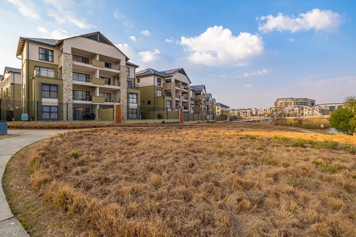 A view of the rear elevation of a modern apartment complex featuring balconies, stone accents, and a paved walkway overlooking a natural landscape.