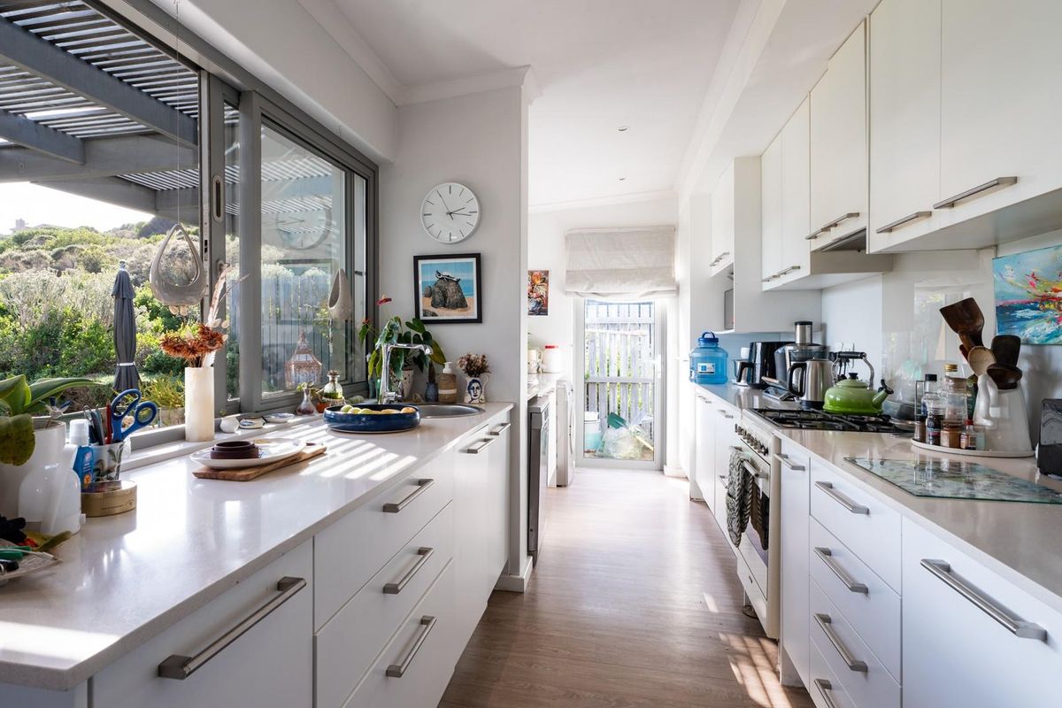 Modern galley-style kitchen featuring white cabinetry, stone benchtops, integrated gas cooktop, and large windows overlooking the outdoor area.