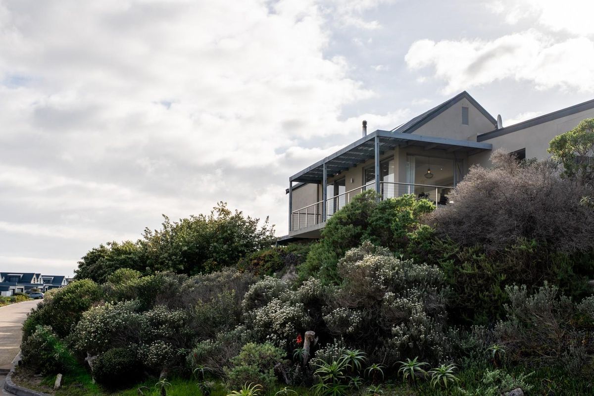 A rear view of a modern house featuring a covered patio with a metal balustrade, set against a landscaped garden and natural hillside.