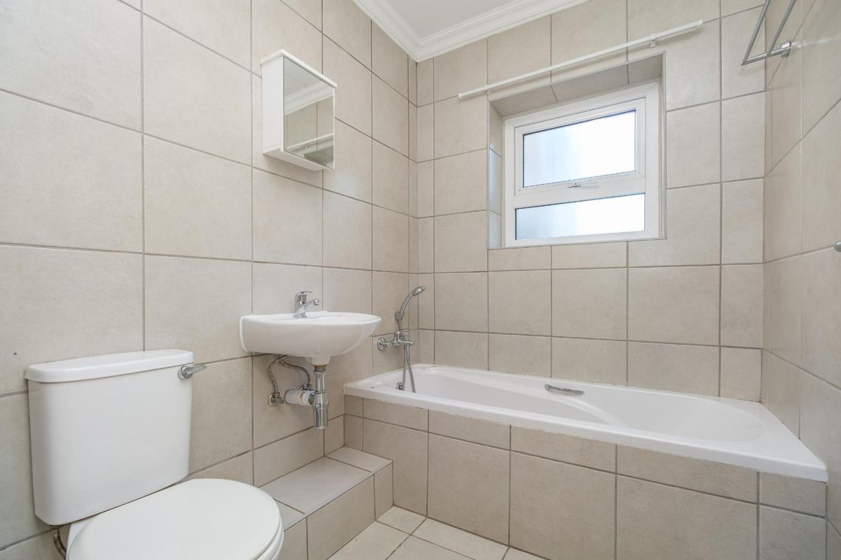 A clean, functional bathroom featuring a bathtub, wall-mounted basin, toilet, and neutral floor-to-ceiling wall tiles.