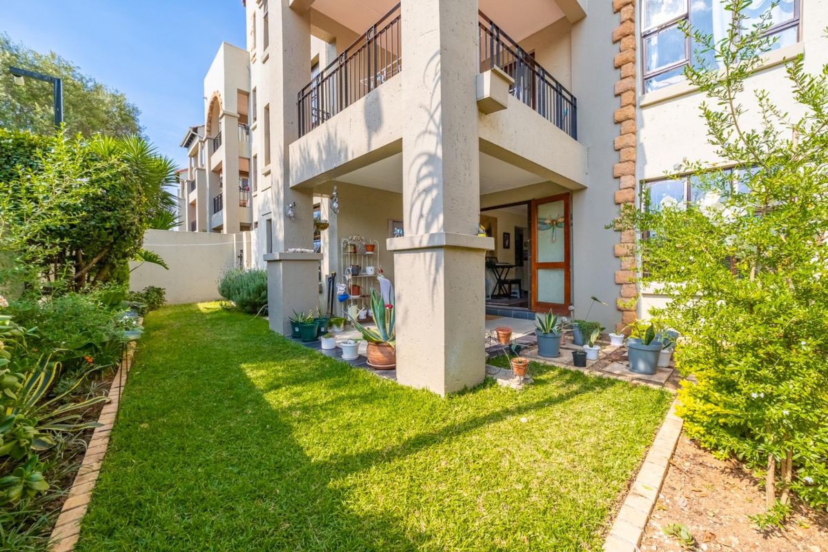 A well-maintained rear exterior view of the apartment featuring a covered patio, balcony with metal railings, and a landscaped garden area.