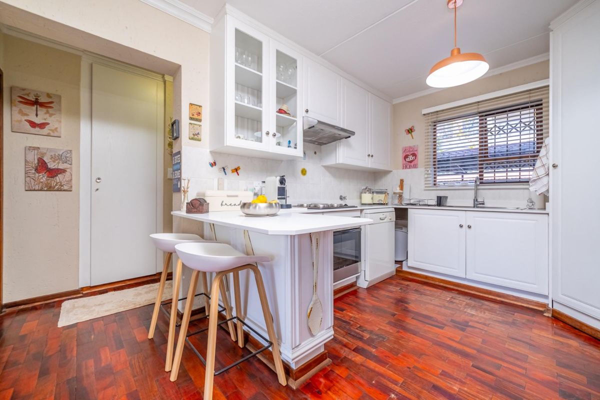 A well-maintained kitchen featuring white cabinetry, a gas hob, oven, extractor fan, and warm wooden parquet flooring with a breakfast counter.