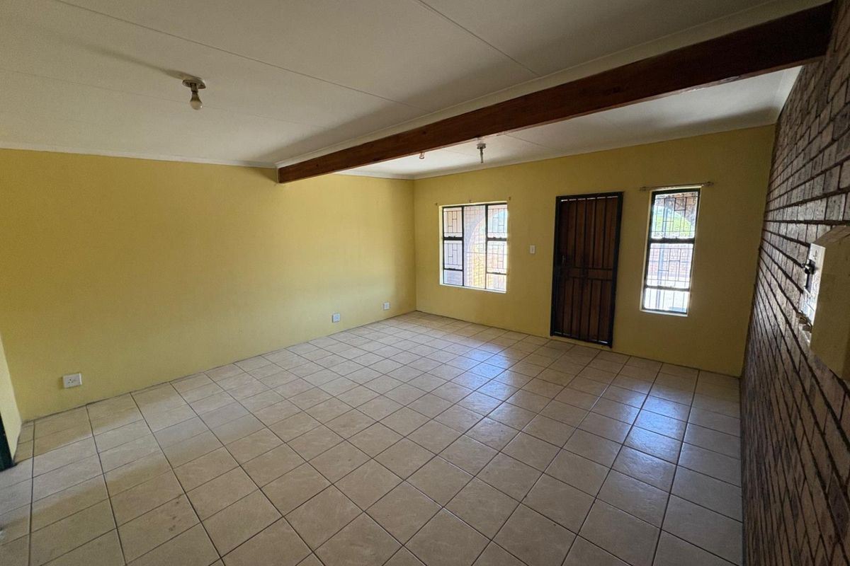 Spacious living area featuring ceramic floor tiles, an exposed timber ceiling beam, and a rustic brick feature wall with secure window and door access.