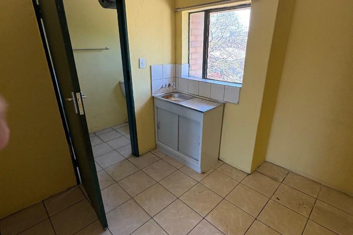 A compact, empty kitchen area featuring tiled flooring, a single stainless steel sink with a tiled splashback, and direct access to an adjacent bathroom.