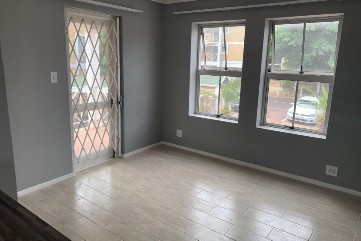 A bright, empty living area featuring neutral tiled flooring, grey walls, and a secure entrance gate with natural light from two windows.
