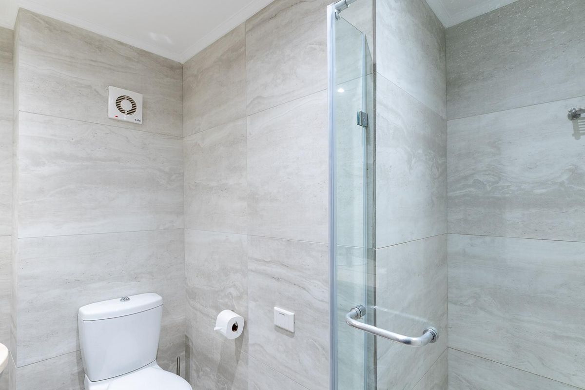A modern bathroom featuring neutral stone-look wall tiles, a glass-enclosed shower, a wall-mounted extractor fan, and a clean, contemporary toilet fixture.