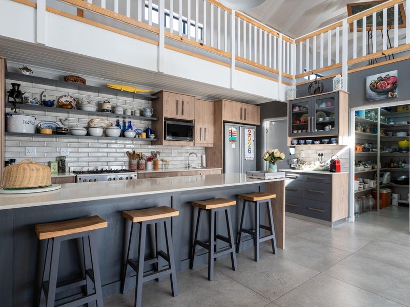 Modern open-plan kitchen featuring a large island with stone countertops, subway tile backsplash, ample cabinetry, and an adjoining scullery under a mezzanine level.