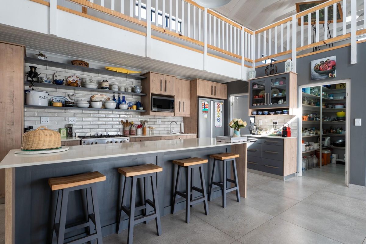Modern open-plan kitchen featuring a large island with stone countertops, subway tile backsplash, ample cabinetry, and an adjoining scullery under a mezzanine level.