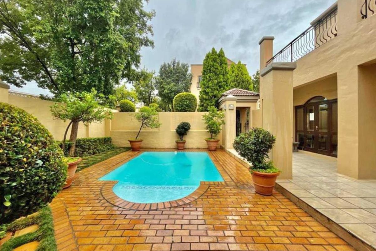 A well-maintained outdoor pool area featuring brick paving, a swimming pool, and a covered patio with a wrought iron balustrade.